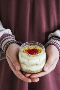Close-up of a person holding a dessert jar filled with layers of cream, biscuit, and topped with a red garnish.