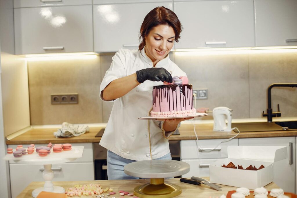 Optimistic woman in apron and latex gloves smiling and putting small sweets on top of cake with chocolate topping while preparing pastry in kitchen